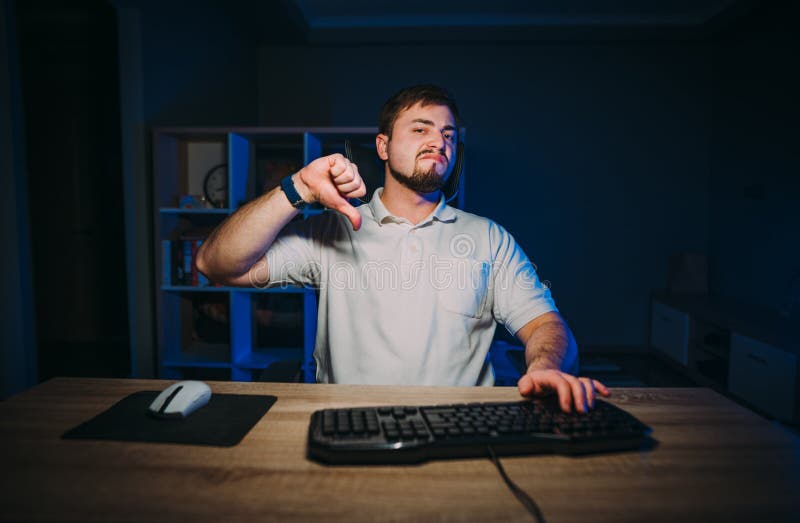 An upset bearded man works at night on a computer at home and shows a dislike gesture. Mirthless freelance programmer shows thumbs royalty free stock image