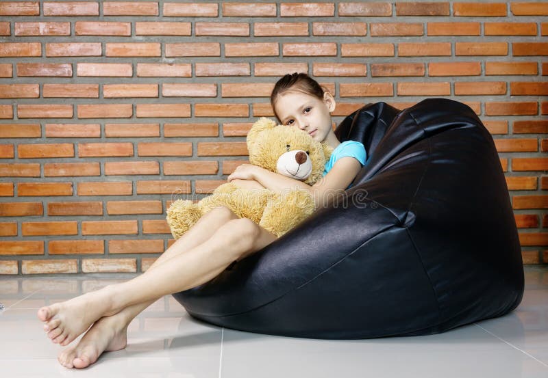 Upset caucasian teen girl sitting in black bean bag chair holding soft teddy bear toy against brick wall. Casual outfit. Child