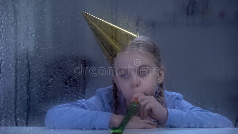 Upset girl in birthday hat blowing party horn on rainy day, celebrating alone, stock photo