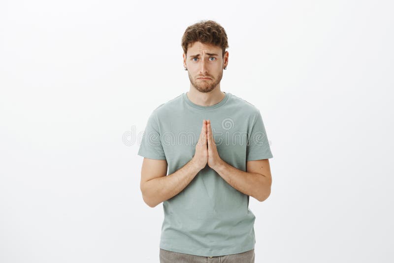 Upset gloomy cute guy with bristle in t-shirt and earrings, holding hands in pray and making sad smile while asking for stock photo Upset gloomy cute guy with bristle in t-shirt and earrings, holding hands in pray and making sad smile while asking for stock photo