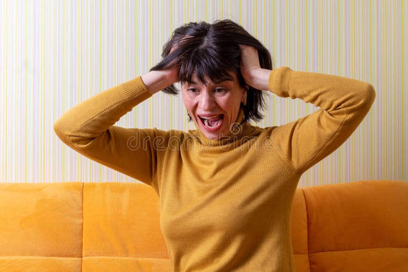 Upset screaming middle aged woman suffering from stress holding her head with her hands on an orange sofa. Middle age crisis. Mental health concept.