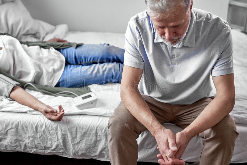 upset senior man sits with her sick ill wife lying on bed royalty free stock photography