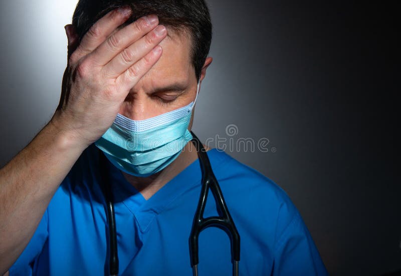 Portrait of a tearful /upset male doctor with hand on head and eyes closed, wearing blue hospital scrubs and a stethoscope with surgical face mask, standing against a dark background. Portrait of a tearful /upset male doctor with hand on head and eyes closed, wearing blue hospital scrubs and a stethoscope with surgical face mask, standing against a dark background.