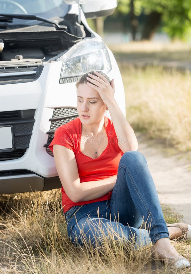 Upset woman sitting on ground and leaning on broken car.