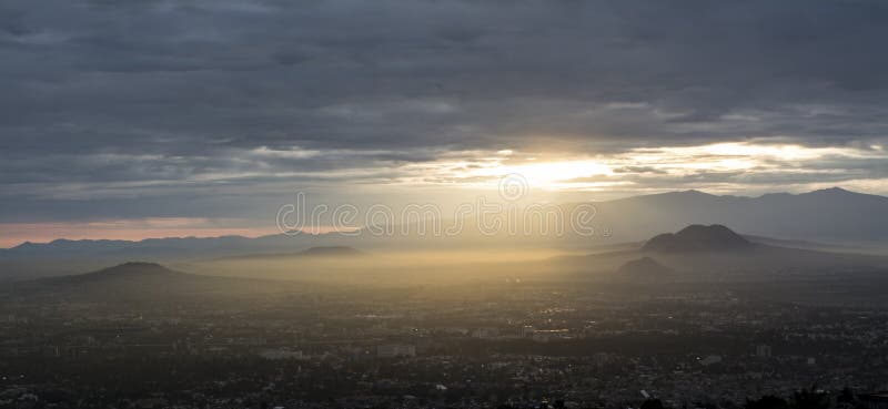 This is a wide, panoramic shot capturing a vast cityscape, likely Mexico City, viewed from a high vantage point, possibly at sunrise or sunset. The foreground shows the densely packed urban area, with buildings and streets visible as a dark, sprawling mass. The middle ground is dominated by a thick layer of golden-hued atmospheric haze or fog that blankets parts of the city and extends towards distant mountains. Sunlight breaks through a dramatic, cloudy sky, creating visible rays that illuminate the hazy layer, giving it a luminous quality. In the background, dark silhouettes of mountains frame the horizon, with some peaks appearing through the mist. The sky above is largely dark and cloudy, contrasting with the bright sun rays. The overall scene evokes a sense of grandeur and the vastness of an urban sprawl nestled within a natural, often hazy, environment. Capturing sunlight stock images, royalty-free photos and pictures
