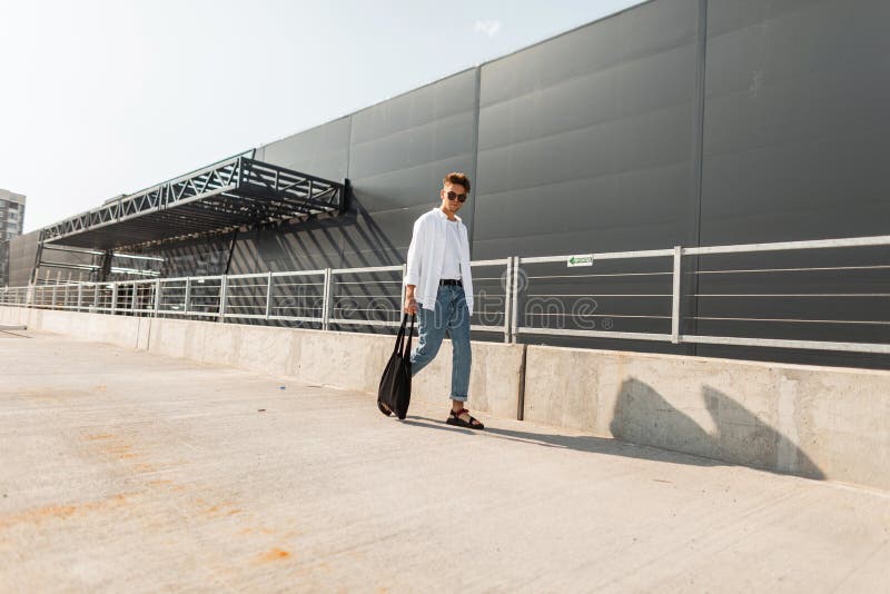 Urban young man hipster in stylish white and denim clothes in trendy sandals with a black cloth bag walks outdoors on a sunny day stock photo Urban young man hipster in stylish white and denim clothes in trendy sandals with a black cloth bag walks outdoors on a sunny day stock photo