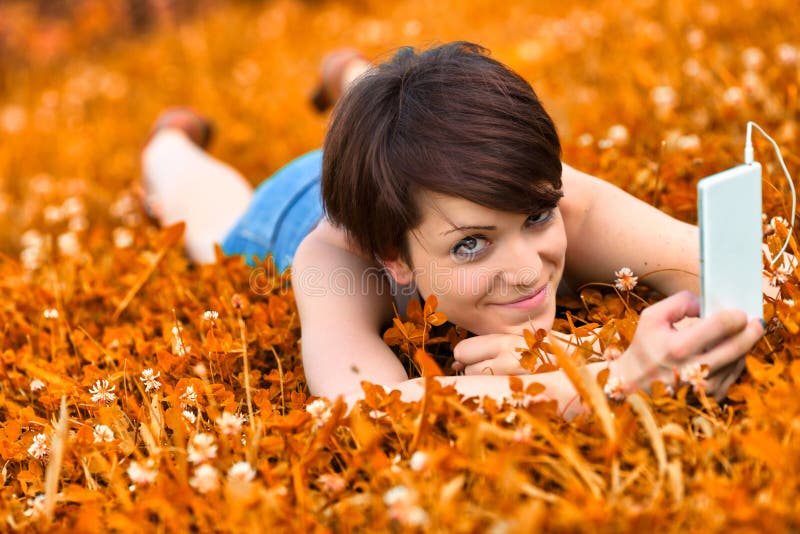 Cute attractive young woman lying in a meadow of orange clover, listening to music on her mobile phone in a head to toe view as she smiles at the camera. Cute attractive young woman lying in a meadow of orange clover, listening to music on her mobile phone in a head to toe view as she smiles at the camera