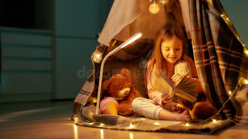 Cute small girl wearing pyjamas sitting on a floor barefoot in a self-made hut made of a plaid, reading a book, smiling and laughing with her teddybear and fancy garlands around the hut in a big room. Cute small girl wearing pyjamas sitting on a floor barefoot in a self-made hut made of a plaid, reading a book, smiling and laughing with her teddybear and fancy garlands around the hut in a big room