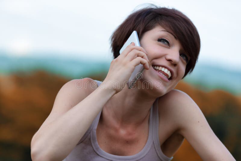 Cute young woman laughing as she chats on her mobile phone outdoors looking up with a beaming smile. Cute young woman laughing as she chats on her mobile phone outdoors looking up with a beaming smile