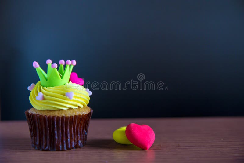 Birthday cup cake with yellow cream and heart for love valentines. green crown on cup cake with cream cheese frosting and 2 hearts on wood floor. Beautiful and delicious cupcake. Birthday cup cake with yellow cream and heart for love valentines. green crown on cup cake with cream cheese frosting and 2 hearts on wood floor. Beautiful and delicious cupcake.