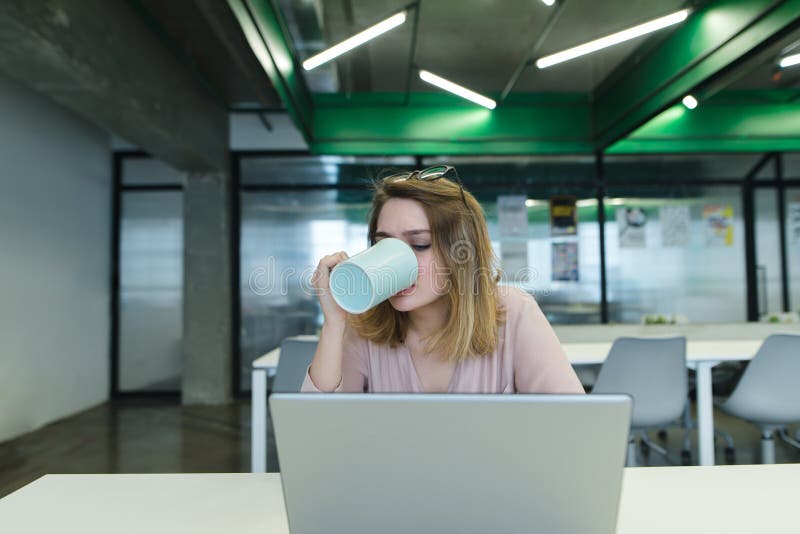 Urzędnik pije kawę podczas gdy pracujący na komputerze Dziewczyna pije gorącego napój od kubka i używa laptop fotografia royalty free Urzędnik pije kawę podczas gdy pracujący na komputerze Dziewczyna pije gorącego napój od kubka i używa laptop fotografia royalty free