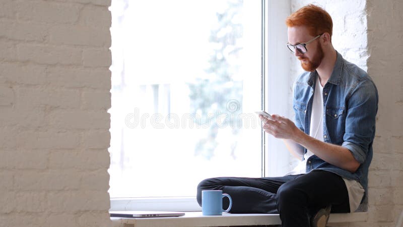 Using Smartphone, Browsing Online by Man Sitting in Window, Beard and Red Hairs
