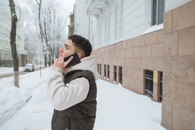 Utomhus- vinterstående för ung stilig man med telefonen Härlig tonåring i hans omslag och väst som poserar på en stadsgata, royaltyfri fotografi Utomhus- vinterstående för ung stilig man med telefonen Härlig tonåring i hans omslag och väst som poserar på en stadsgata, royaltyfri fotografi