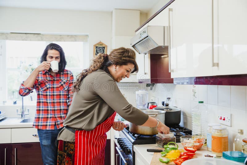 Mid adult men is relaxing in the kitchen talking to his mother while she prepares a curry on the cooker. Mid adult men is relaxing in the kitchen talking to his mother while she prepares a curry on the cooker.