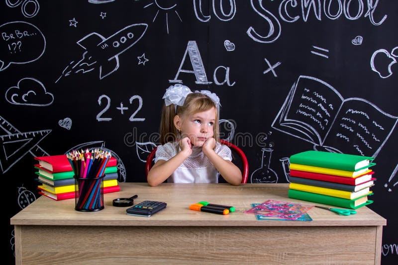 Uttråkat och drömma skolflickasammanträde på skrivbordet med böcker, skolatillförsel som rymmer hennes båda händer över cheecksna arkivbilder Uttråkat och drömma skolflickasammanträde på skrivbordet med böcker, skolatillförsel som rymmer hennes båda händer över cheecksna arkivbilder
