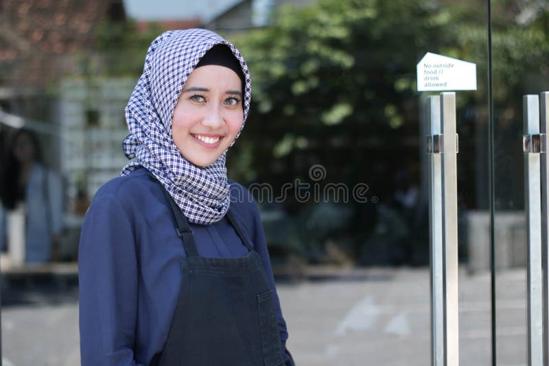 Uśmiechać się Hijab Arabskiej kobiety wita kostiumera z fartuchem pozuje przed szklanym drzwi fotografia royalty free Uśmiechać się Hijab Arabskiej kobiety wita kostiumera z fartuchem pozuje przed szklanym drzwi fotografia royalty free