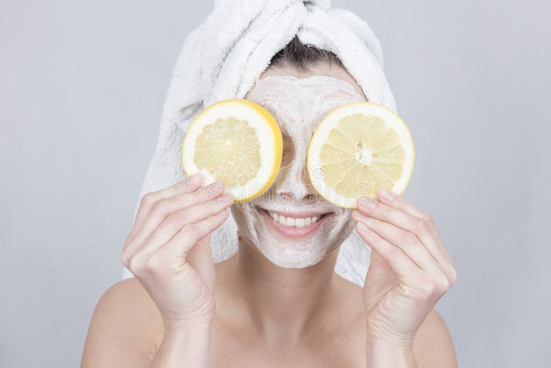 Uśmiechnięta brunetki kobieta trzyma dwa plasterek cytryna przed jej twarzą kobieta z nawilżać twarzową maskę obrazy stock Uśmiechnięta brunetki kobieta trzyma dwa plasterek cytryna przed jej twarzą kobieta z nawilżać twarzową maskę obrazy stock