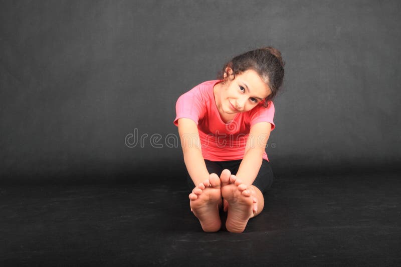 Smiling pretty girl - barefoot brunette schoolkid dressed in pink t-shirt, exercising yoga pose seated forward fold or paschimottanasana with hands reaching soles of bare feet. Active childhood concept. Smiling pretty girl - barefoot brunette schoolkid dressed in pink t-shirt, exercising yoga pose seated forward fold or paschimottanasana with hands reaching soles of bare feet. Active childhood concept.