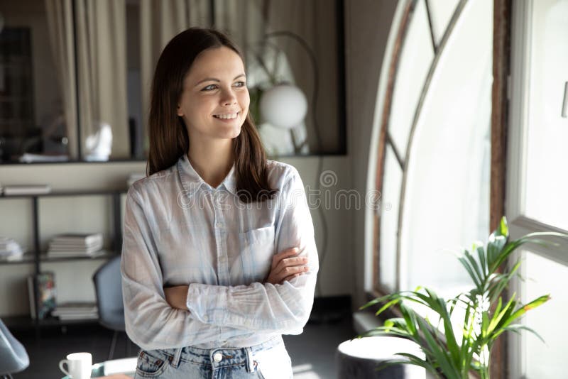 Smiling young caucasian female employee look in window distance visualizing or dreaming at workplace, happy millennial woman worker thinking pondering, planning career success, business vision concept. Smiling young caucasian female employee look in window distance visualizing or dreaming at workplace, happy millennial woman worker thinking pondering, planning career success, business vision concept