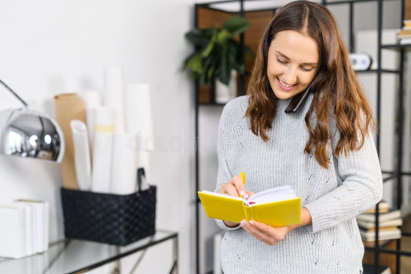 Cheerful woman has phone conversation and writes down into notebook, standing in the office, multitasking female office employee. Cheerful woman has phone conversation and writes down into notebook, standing in the office, multitasking female office employee