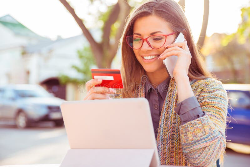 Uśmiechnięta kobieta siedzi outdoors opowiadać na telefonie komórkowym robi online zapłacie na jej pastylka komputerze obraz stock Uśmiechnięta kobieta siedzi outdoors opowiadać na telefonie komórkowym robi online zapłacie na jej pastylka komputerze obraz stock