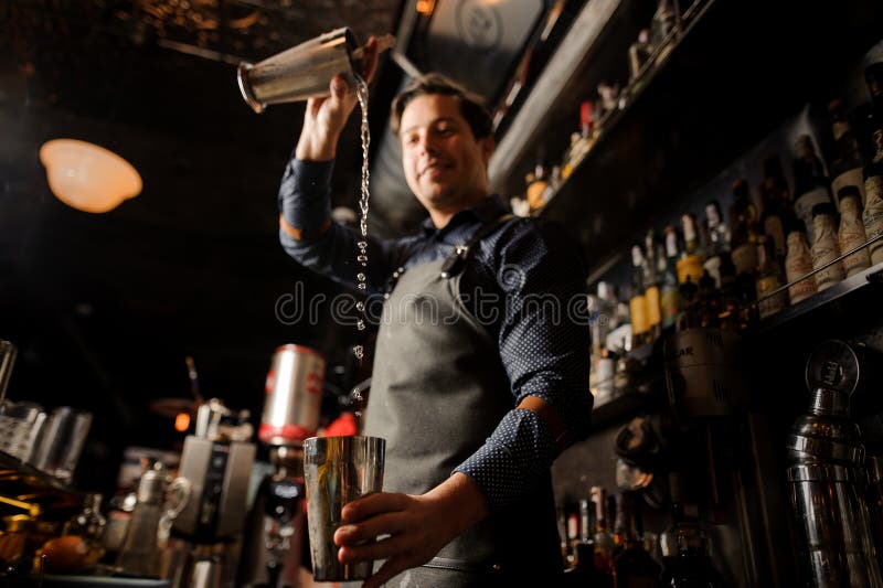 Uśmiechnięty barman nalewa alkoholicznego napój od jeden metalu szkła w inny fotografia stock Uśmiechnięty barman nalewa alkoholicznego napój od jeden metalu szkła w inny fotografia stock