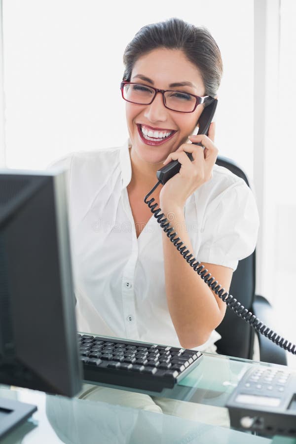 Uśmiechnięty bizneswomanu obsiadanie przy jej biurkiem opowiada na telefonie obraz royalty free Uśmiechnięty bizneswomanu obsiadanie przy jej biurkiem opowiada na telefonie obraz royalty free