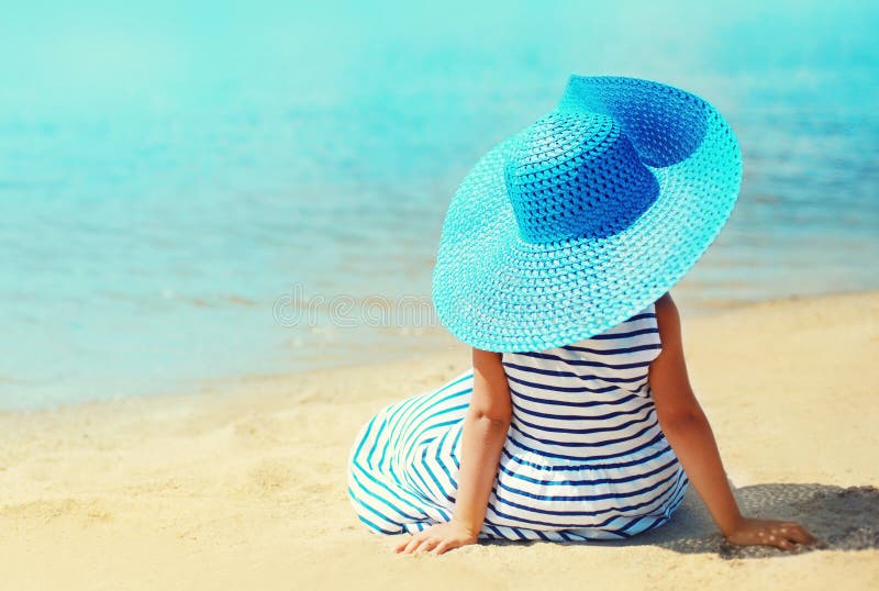 Vacaciones de verano y concepto de las vacaciones - niña en el vestido rayado, sombrero de paja disfrutando de sentarse en la pla imagen de archivo libre de regalías Vacaciones de verano y concepto de las vacaciones - niña en el vestido rayado, sombrero de paja disfrutando de sentarse en la pla imagen de archivo libre de regalías