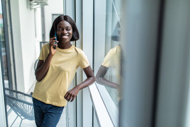 Vacker amerikansk afro-flicka som pratar på mobiltelefonen och leende medan han stod nära fönstret fotografering för bildbyråer