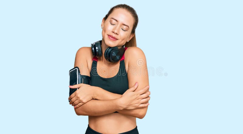 Vacker, blond kvinna med gym kläder och hörlurar som kramar sig själv glad och positiv och leende självsäker fotografering för bildbyråer