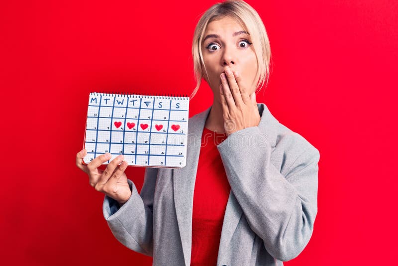 Vacker, blond kvinna som kontrollerar menstruationstiden, kalender över röd bakgrund som täcker munnen med handchockad och royaltyfria bilder