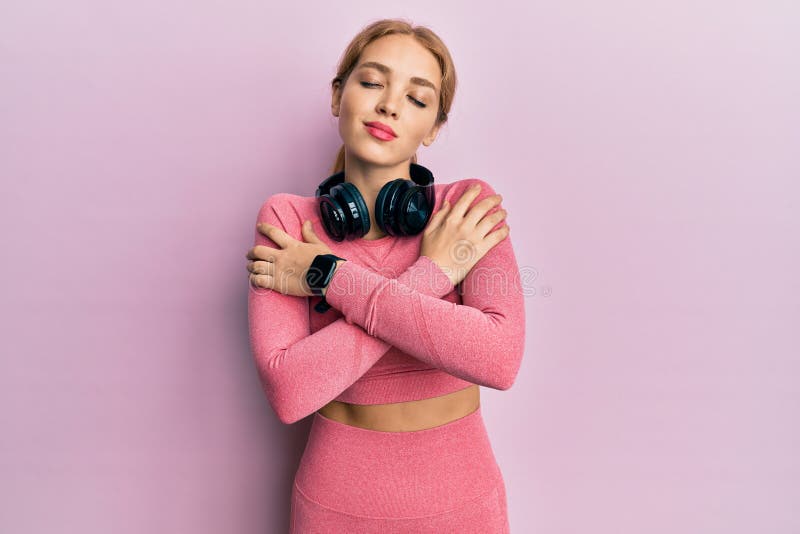 Vacker blonde kaukasisk kvinna som bär gym kläder och använder hörlurar som kramar sig själv, glad och positiv, leende självsäker royaltyfri fotografi