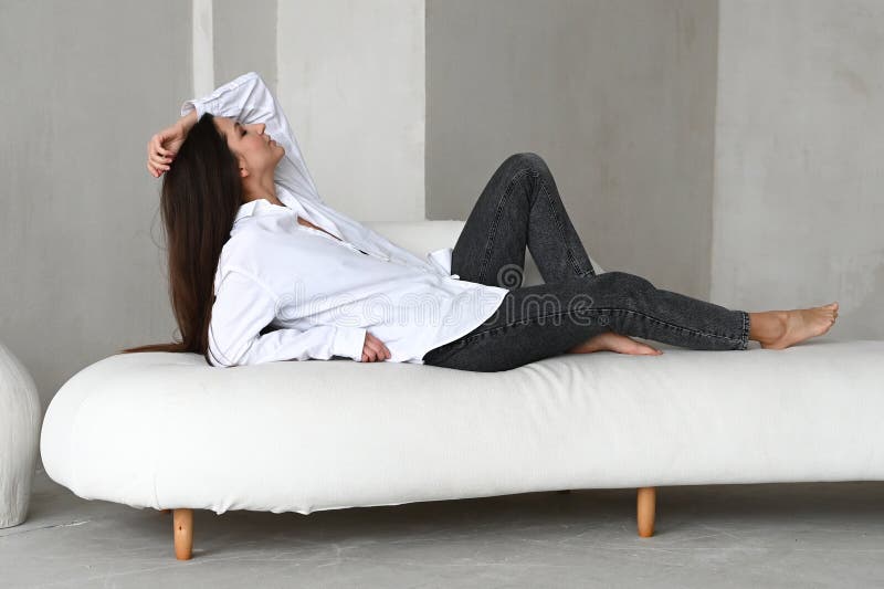 Vacker brunette som pockar i studio medan man ligger på en vit sofa. vacker brunette ser ut att bli tanklös fotografering för bildbyråer