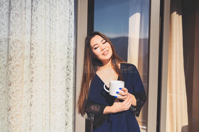 Vacker, leende kvinna som njuter av kaffe eller te på solig balkong. ung, lycklig dam i blå badrock, kyld arkivbilder