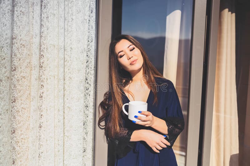 Vacker, leende kvinna som njuter av kaffe eller te på solig balkong. ung, lycklig dam i blå badrock, kyld royaltyfri foto