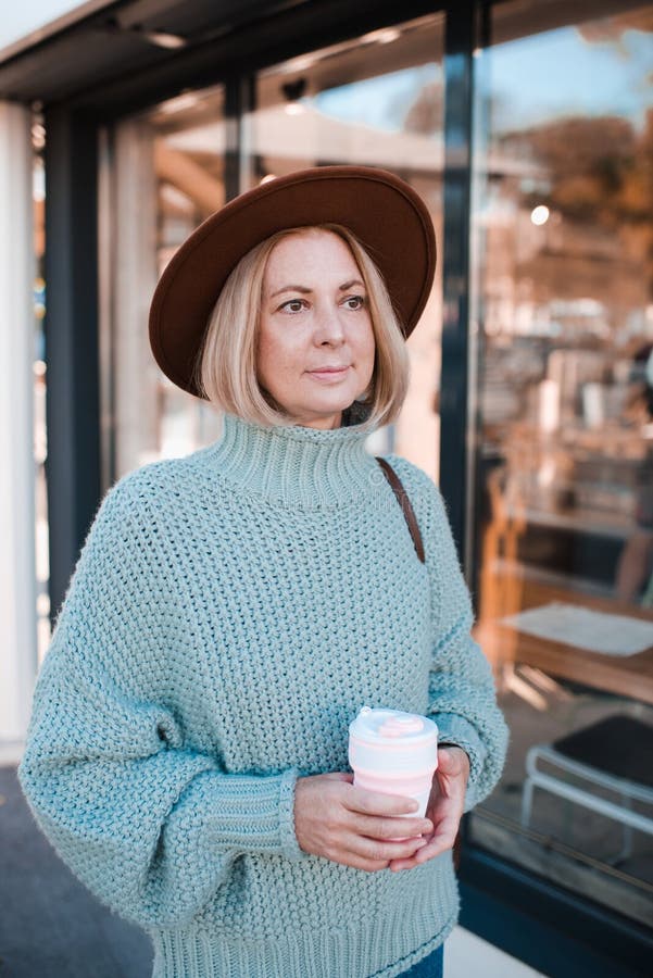 Beautiful mature woman 50-55 year old drink coffee in paper cup wear knit sweater and felt hat in cafe outdoors. Autumn season. Healthy aged blonde female. Happiness. Beautiful mature woman 50-55 year old drink coffee in paper cup wear knit sweater and felt hat in cafe outdoors. Autumn season. Healthy aged blonde female. Happiness