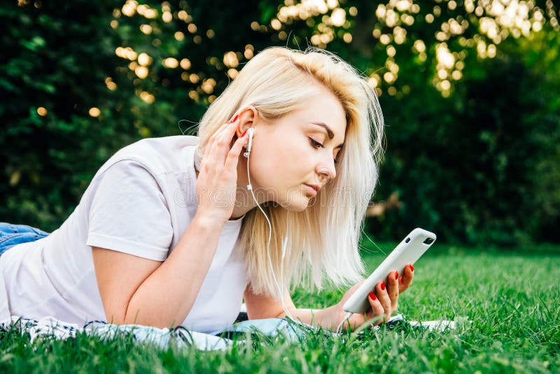 Vackra, blonda kvinnan i hörlurarna och med en smartphone i händerna som lyssnar på musik som ligger på gräs utomhus i parken Pla royaltyfri fotografi