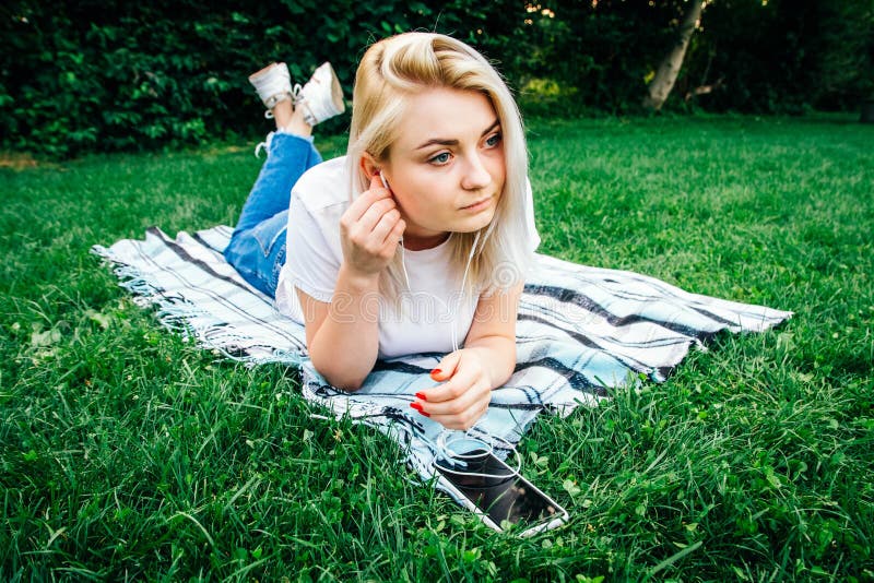 Vackra, blonda kvinnan i hörlurarna och med en smartphone i händerna som lyssnar på musik som ligger på gräs utomhus i parken Pla royaltyfria bilder