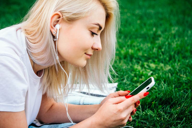 Vackra, blonda kvinnan i hörlurarna och med en smartphone i händerna som lyssnar på musik som ligger på gräs utomhus i parken Pla royaltyfria foton