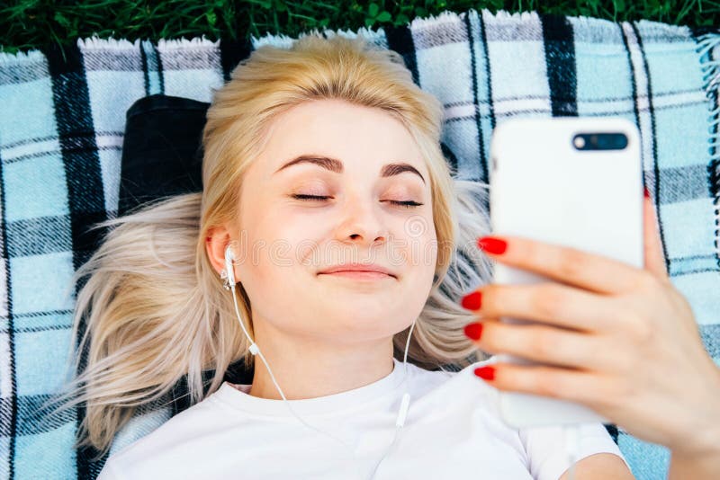 Vackra, blonda kvinnan i hörlurarna och med en smartphone i händerna som lyssnar på musik som ligger på gräs utomhus i parken Övr royaltyfri fotografi