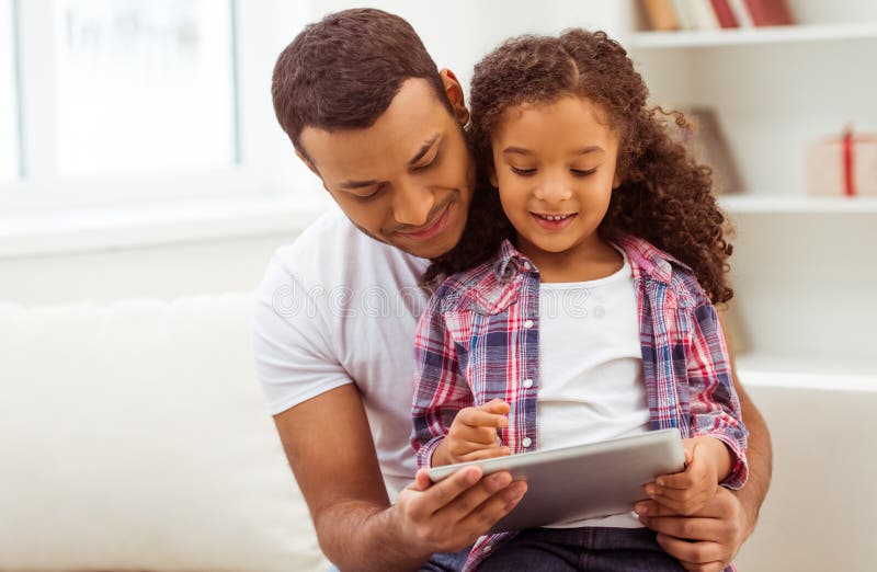 Cute little Afro-American girl in casual clothes sitting on her father's knees. Both using a tablet and smiling. Cute little Afro-American girl in casual clothes sitting on her father's knees. Both using a tablet and smiling.