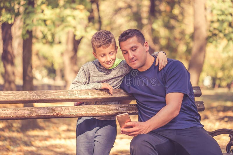 Vader en zoon die een onderbreking na training nemen en op iets op smartphone in het park letten stock foto Vader en zoon die een onderbreking na training nemen en op iets op smartphone in het park letten stock foto