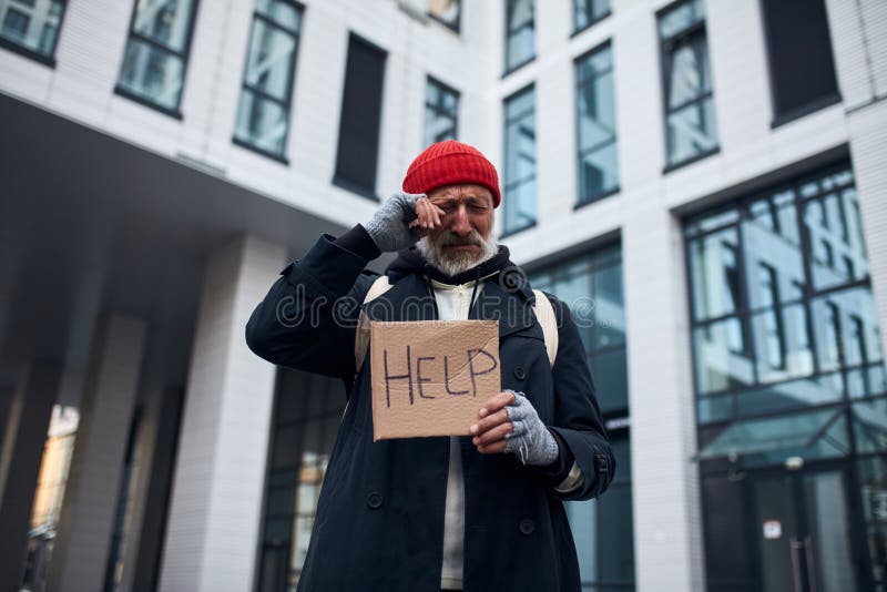 Vagrant ber om hjälp i stadens centrum med hjälpskylt. Torkmannen som håller hjälplinje tillverkad av papp, står i stadens centrum, en vacker affärsbyggnad bakom honom
