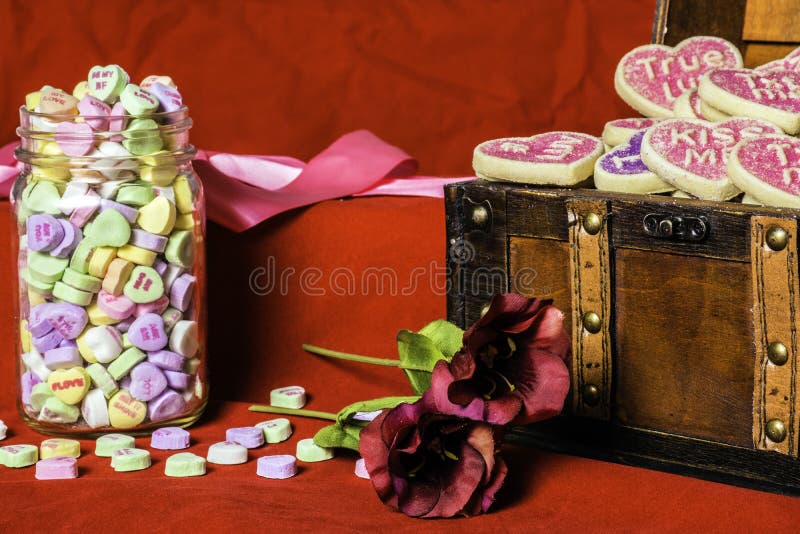 Wood chest full of valentine conversation cookies and glass jar full of conversation candy hearts on red background with two red flowers and pink ribbon. Wood chest full of valentine conversation cookies and glass jar full of conversation candy hearts on red background with two red flowers and pink ribbon