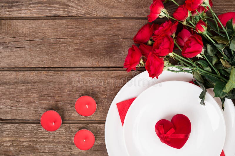 Valentine`s Day dinner table setting with red ribbon, roses, knife and fork ring over oak background.