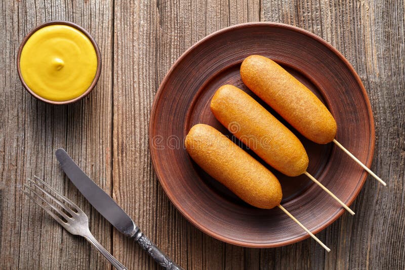 Corn dog traditional American corndog street junk food deep fried hotdog meat sausage snack coated in a thick layer of cornmeal batter on stick unhealthy eating on rustic wooden background table. Corn dog traditional American corndog street junk food deep fried hotdog meat sausage snack coated in a thick layer of cornmeal batter on stick unhealthy eating on rustic wooden background table.