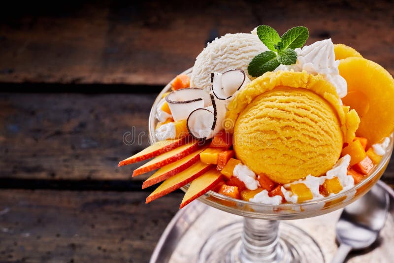 High Angle Close Up View of Sundae Made with Scoops of Vanilla and Peach Flavored Ice Cream, Slices of Fresh Fruit, Whipped Cream and Mint Garnish Served in Glass Dish on Wood Table with Copy Space. High Angle Close Up View of Sundae Made with Scoops of Vanilla and Peach Flavored Ice Cream, Slices of Fresh Fruit, Whipped Cream and Mint Garnish Served in Glass Dish on Wood Table with Copy Space