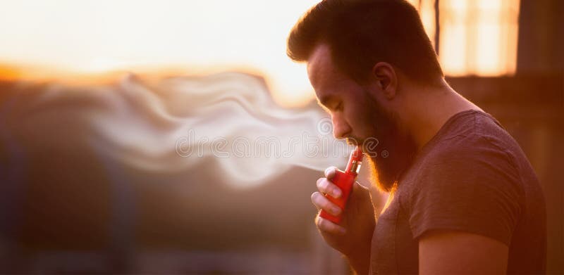 Vaping, молодой человек с бородой, производит предпосылку неба захода солнца пара, место для текста стоковое фото rf Vaping, молодой человек с бородой, производит предпосылку неба захода солнца пара, место для текста стоковое фото rf