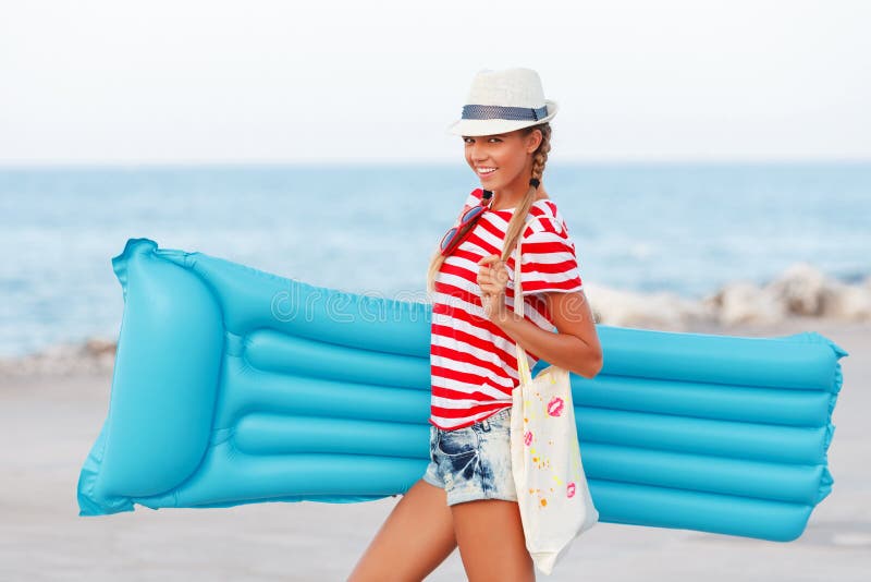 Vare el sombrero feliz y que lleva de la mujer de la playa con el colchón azul que se divierte el verano durante vacaciones de lo fotos de archivo libres de regalías Vare el sombrero feliz y que lleva de la mujer de la playa con el colchón azul que se divierte el verano durante vacaciones de lo fotos de archivo libres de regalías