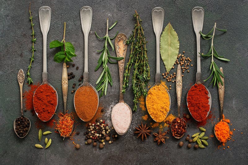 A variety of spices in spoons and herbs on a dark rustic background. Top view, flat lay